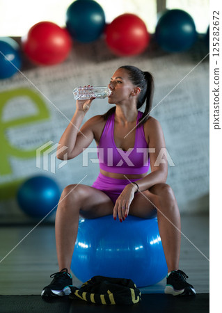 A fit woman takes a well deserved break in the gym, sipping water from her bottle after an intense workout session A fit woman takes a well deserved break in the gym, sipping water from her bottle after an intense workout session 125282672