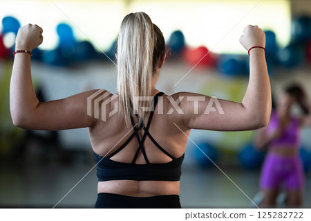 A fitness woman posing in the gym, proudly showing off her biceps and showcasing her strength and dedication to fitness. 125282722