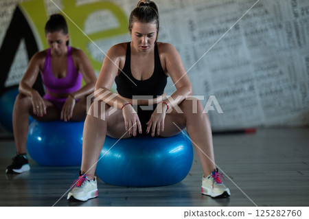 Two fitness women resting and recovering in the gym after an intense workout session, showcasing strength, endurance, and dedication to their fitness journey. Two fitness women resting and recovering in the gym after an intense workout session, showcasing strength, endurance, and dedication to their fitness journey. 125282760