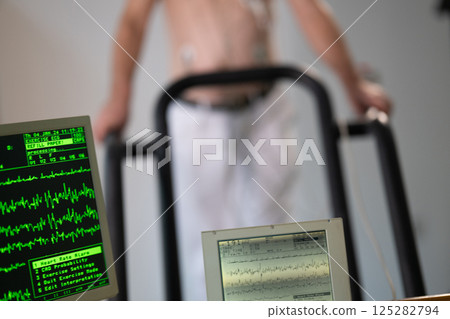 A middle-aged man is undergoing a medical stress test on a treadmill, connected to ECG electrodes for heart monitoring in a clinical environment 125282794