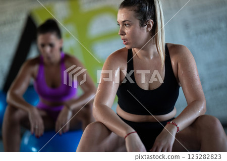 Two fitness women resting and recovering in the gym after an intense workout session, showcasing strength, endurance, and dedication to their fitness journey. Two fitness women resting and recovering in the gym after an intense workout session, showcasing strength, endurance, and dedication to their fitness journey. 125282823