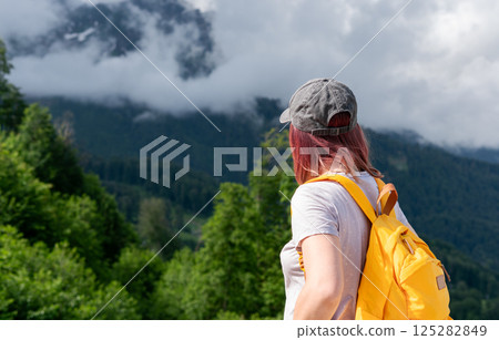 Woman hiker with red hair yellow backpack and cap views mountain landscape surrounded by lush greenery and cloudy sky, moment of reflection and adventure in nature, copy space, hiking Woman hiker with red hair yellow backpack and cap views mountain landscape surrounded by lush greenery and cloudy sky, moment of reflection and adventure in nature, copy space, hiking 125282849