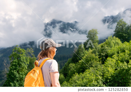 Woman hiker with yellow backpack and cap looks at a foggy mountain landscape surrounded by lush greenery, adventure and tranquility in nature setting, hiking and active healthy lifestyle , copy space Woman hiker with yellow backpack and cap looks at a foggy mountain landscape surrounded by lush greenery, adventure and tranquility in nature setting, hiking and active healthy lifestyle , copy space 125282850