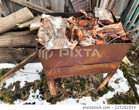 Rusted metal fire pit filled with birch logs and wood pieces. Surrounded by logs and patches of snow, the scene highlights a rustic outdoor setting, perfect for backyard fires and winter warmth Rusted metal fire pit filled with birch logs and wood pieces. Surrounded by logs and patches of snow, the scene highlights a rustic outdoor setting, perfect for backyard fires and winter warmth 125282854