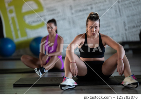 Two fitness women resting and recovering in the gym after an intense workout session, showcasing strength, endurance, and dedication to their fitness journey. Two fitness women resting and recovering in the gym after an intense workout session, showcasing strength, endurance, and dedication to their fitness journey. 125282876