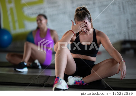 Two fitness women resting and recovering in the gym after an intense workout session, showcasing strength, endurance, and dedication to their fitness journey. Two fitness women resting and recovering in the gym after an intense workout session, showcasing strength, endurance, and dedication to their fitness journey. 125282889