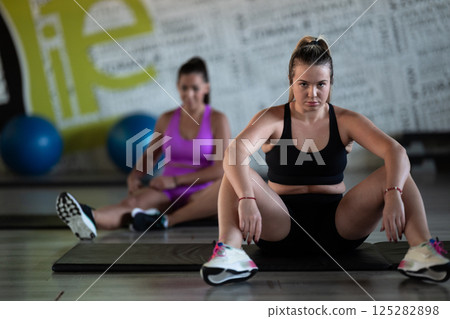 Two fitness women resting and recovering in the gym after an intense workout session, showcasing strength, endurance, and dedication to their fitness journey. Two fitness women resting and recovering in the gym after an intense workout session, showcasing strength, endurance, and dedication to their fitness journey. 125282898