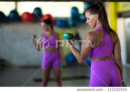 A focused and determined woman performs a strength building dumbbell workout at the gym, showcasing her athletic physique and dedication to fitness. 125282919