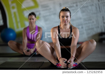 Two fitness women stretch together in the gym, supporting each other in their journey towards flexibility and wellness. Two fitness women stretch together in the gym, supporting each other in their journey towards flexibility and wellness. 125282958