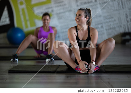 Two fitness women stretch together in the gym, supporting each other in their journey towards flexibility and wellness. Two fitness women stretch together in the gym, supporting each other in their journey towards flexibility and wellness. 125282985