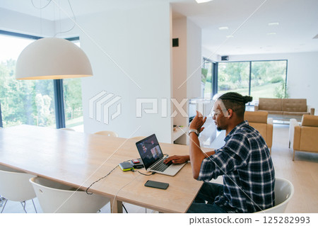 African American influencer in glasses sitting at a table in a modern living room, using a laptop for business video chat, conversation with friends and entertainment. 125282993