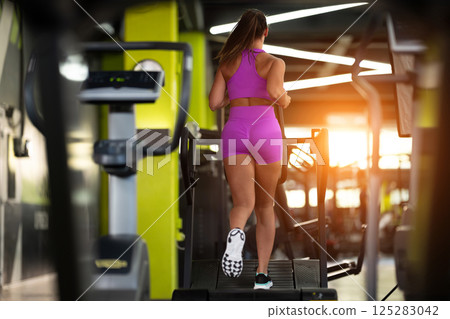 Woman Running on Treadmill in Gym.A fit woman wearing a purple sports outfit is running on a treadmill in a gym. She has a focused yet relaxed expression, with gym equipment and a warm light in the Woman Running on Treadmill in Gym.A fit woman wearing a purple sports outfit is running on a treadmill in a gym. She has a focused yet relaxed expression, with gym equipment and a warm light in the 125283042