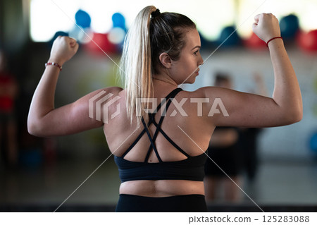 A fitness woman posing in the gym, proudly showing off her biceps and showcasing her strength and dedication to fitness. A fitness woman posing in the gym, proudly showing off her biceps and showcasing her strength and dedication to fitness. 125283088