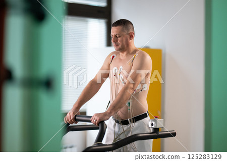 A middle-aged man is undergoing a medical stress test on a treadmill, connected to ECG electrodes for heart monitoring in a clinical environment A middle-aged man is undergoing a medical stress test on a treadmill, connected to ECG electrodes for heart monitoring in a clinical environment 125283129