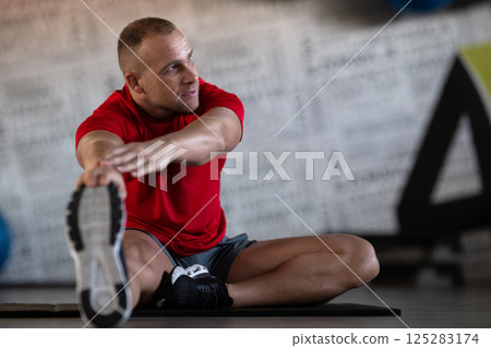 A fitness man stretches in the gym after an intense workout, focusing on recovery and flexibility. A fitness man stretches in the gym after an intense workout, focusing on recovery and flexibility. 125283174