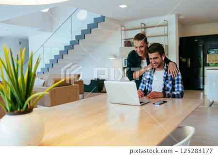 A young married couple is talking to parents, family and friends on a video call via a laptop while sitting in the living room of their modern house. A young married couple is talking to parents, family and friends on a video call via a laptop while sitting in the living room of their modern house. 125283177