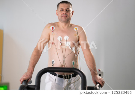 A middle-aged man is undergoing a medical stress test on a treadmill, connected to ECG electrodes for heart monitoring in a clinical environment A middle-aged man is undergoing a medical stress test on a treadmill, connected to ECG electrodes for heart monitoring in a clinical environment 125283495