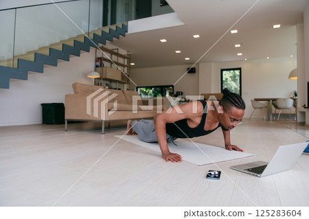 Training At Home. Sporty african american man doing the training while watching an online tutorial on a laptop, exercising in the living room, free space.  125283604