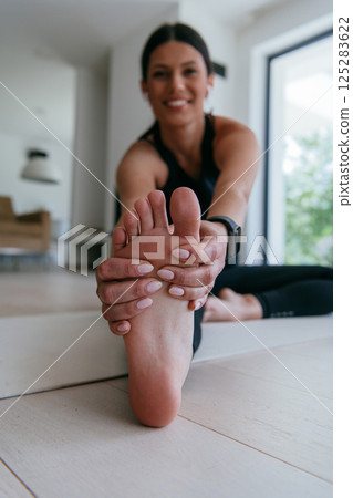 A young woman in sportswear stretching her legs during online training in the living room of her home. 125283622
