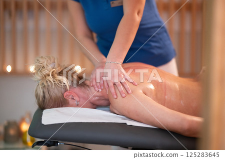 Relaxing Back Massage in a Spa Setting. A professional massage therapist in a blue uniform provides a relaxing back massage to a client lying on a massage table. Relaxing Back Massage in a Spa Setting. A professional massage therapist in a blue uniform provides a relaxing back massage to a client lying on a massage table. 125283645