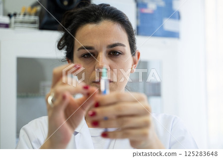 A focused nurse in a white lab coat carefully prepares a syringe for an injection in a professional healthcare environment. 125283648