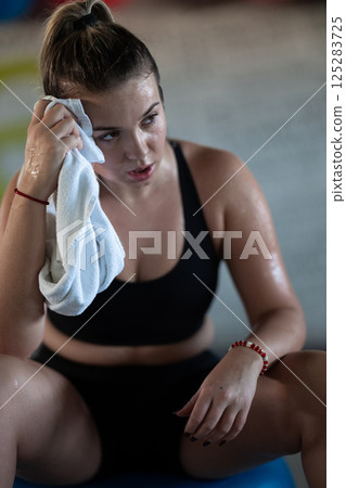 A beautiful and fit woman sitting on a gym ball with a towel, resting and recovering after an intense workout. 125283725