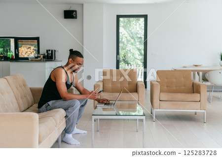 African American man in glasses sitting at a table in a modern living room, using a laptop for business video chat, conversation with friends and entertainment 125283728