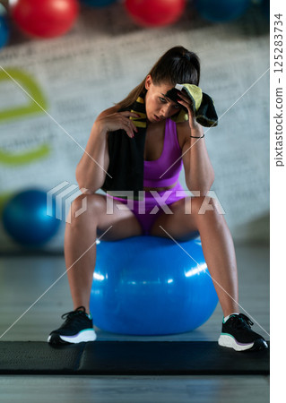 A fit woman takes a well deserved break in the gym, sipping water from her bottle after an intense workout session A fit woman takes a well deserved break in the gym, sipping water from her bottle after an intense workout session 125283734