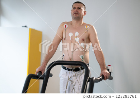 A middle-aged man is undergoing a medical stress test on a treadmill, connected to ECG electrodes for heart monitoring in a clinical environment 125283761