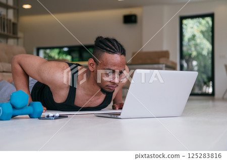 Training At Home. Sporty african american man doing the training while watching an online tutorial on a laptop, exercising in the living room, free space.  125283816