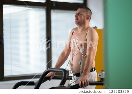 A middle-aged man is undergoing a medical stress test on a treadmill, connected to ECG electrodes for heart monitoring in a clinical environment A middle-aged man is undergoing a medical stress test on a treadmill, connected to ECG electrodes for heart monitoring in a clinical environment 125283859