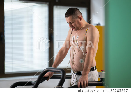A middle-aged man is undergoing a medical stress test on a treadmill, connected to ECG electrodes for heart monitoring in a clinical environment 125283867