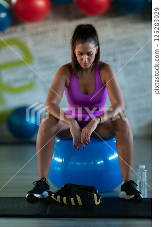 A fit woman takes a well deserved break in the gym, sipping water from her bottle after an intense workout session A fit woman takes a well deserved break in the gym, sipping water from her bottle after an intense workout session 125283929