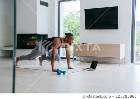 Training At Home. Sporty african american man doing the training while watching an online tutorial on a laptop, exercising in the living room, free space. Training At Home. Sporty african american man doing the training while watching an online tutorial on a laptop, exercising in the living room, free space. 125283965