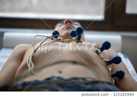 A female doctor performs an ECG on a young man in a hospital room, carefully monitoring his heart health during a routine checkup. 125283999