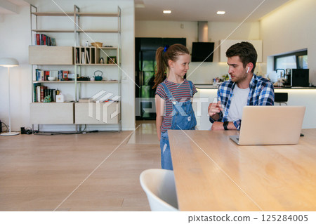 Work from home. Daughter interrupting her father while he is having a business online conversation on his laptop while sitting in the modern living room. 125284005