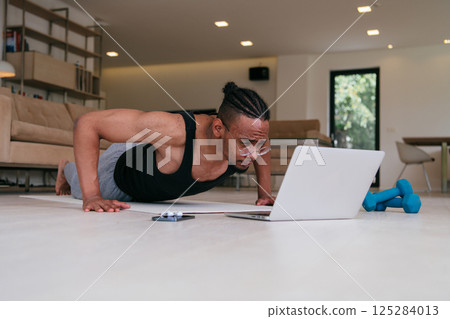 Training At Home. Sporty african american man doing the training while watching an online tutorial on a laptop, exercising in the living room, free space.  125284013