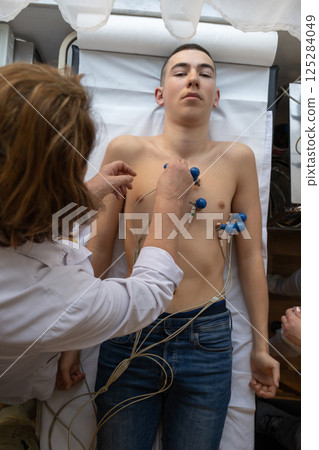 A female doctor performs an ECG on a young man in a hospital room, carefully monitoring his heart health during a routine checkup. A female doctor performs an ECG on a young man in a hospital room, carefully monitoring his heart health during a routine checkup. 125284049
