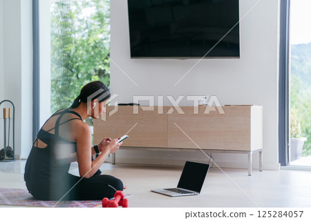 Young woman in sportswear using laptop and smartphone for online training in living room at home. 125284057