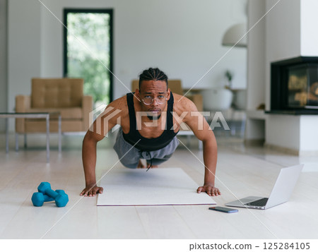 Training At Home. Sporty man doing the training while watching an online tutorial on a laptop, exercising in the living room, free space. 125284105