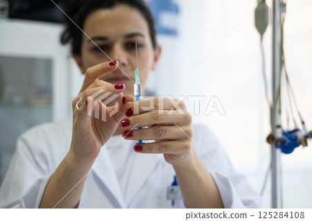 A focused nurse in a white lab coat carefully prepares a syringe for an injection in a professional healthcare environment. 125284108
