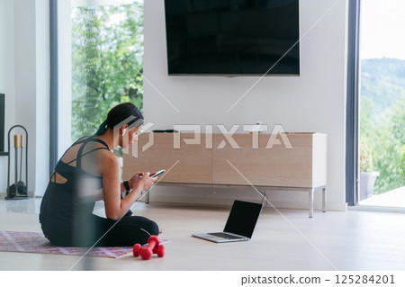 Young woman in sportswear using laptop and smartphone for online training in living room at home. 125284201