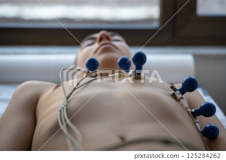 A female doctor performs an ECG on a young man in a hospital room, carefully monitoring his heart health during a routine checkup. A female doctor performs an ECG on a young man in a hospital room, carefully monitoring his heart health during a routine checkup. 125284262