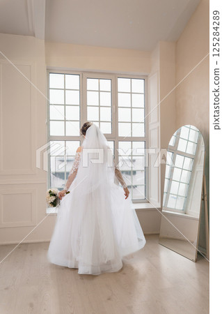 Elegant bride standing with back towards camera in a bright room with large windows, holding a bouquet, preparing for wedding ceremony 125284289