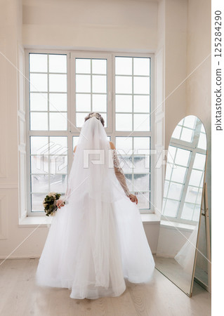 Back view of a Caucasian bride in a white wedding dress and veil standing by a window with a mirror and bouquet Back view of a Caucasian bride in a white wedding dress and veil standing by a window with a mirror and bouquet 125284290