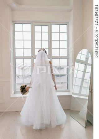 Elegant bride in a white lace wedding dress with a long veil stands with her back to the camera near a window looking to the city Elegant bride in a white lace wedding dress with a long veil stands with her back to the camera near a window looking to the city 125284291