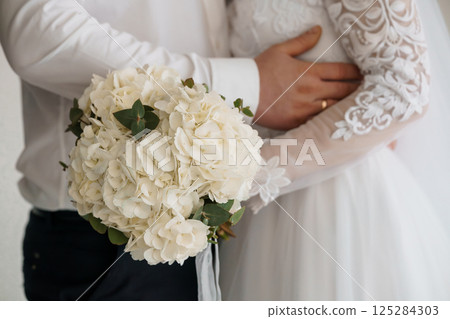 Intimate Wedding Moment: Groom Embracing Bride Holding White Hydrangea Bouquet with Lace Sleeve Details and Elegant Attire, symbolizing love and commitment Intimate Wedding Moment: Groom Embracing Bride Holding White Hydrangea Bouquet with Lace Sleeve Details and Elegant Attire, symbolizing love and commitment 125284303