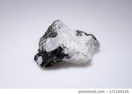 Raw moonstone specimen with black inclusions against a plain white background, showcasing its natural texture and iridescent sheen 125284326