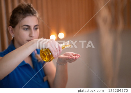 A professional massage therapist pours aromatic oil into their hands, preparing for a relaxing and rejuvenating massage session in a serene spa setting. 125284370