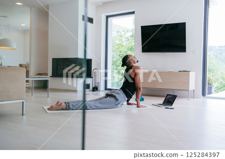 Training At Home. Sporty african american man doing the training while watching an online tutorial on a laptop, exercising in the living room, free space.  125284397